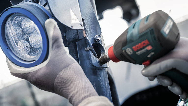 Person wearing safety equipment uses a power tool to fasten a metal bracket near a light.