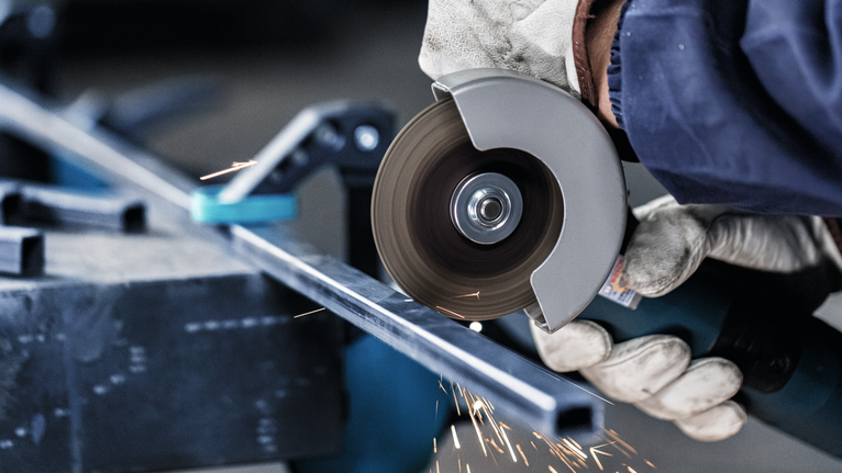 PRO Metal Cutting Disc, 100 x 2.5 x 16 mm A worker wearing safety equipment cuts a metal bar with an angle grinder.