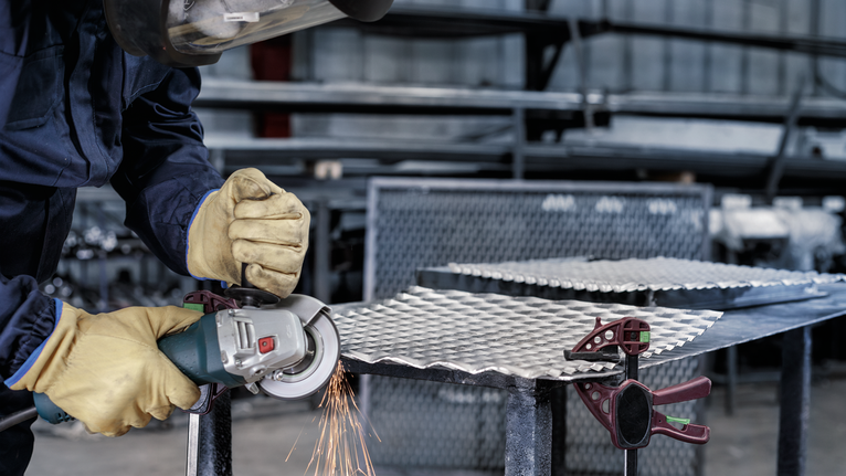 A person wearing safety equipment grinds metal sheeting on a workbench.