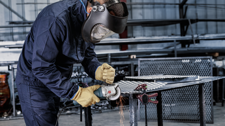 A person wearing safety equipment grinds a metal sheet, producing sparks.