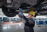 A person wearing safety equipment uses a power wrench to fasten bolts under a vehicle chassis.