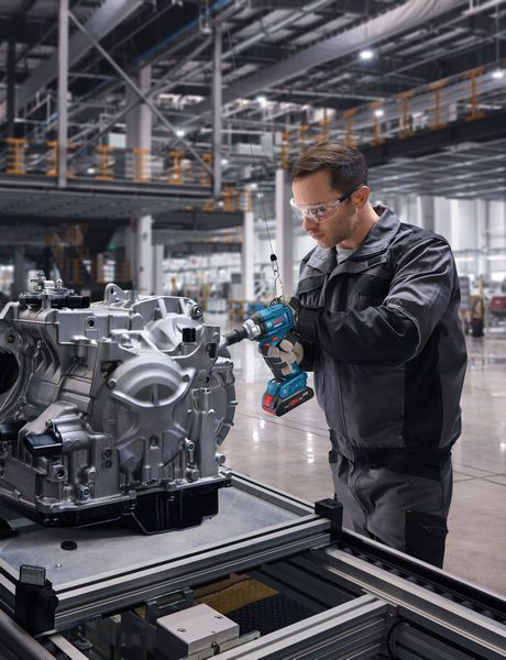 A person wearing safety equipment uses a cordless impact wrench on an engine assembly.