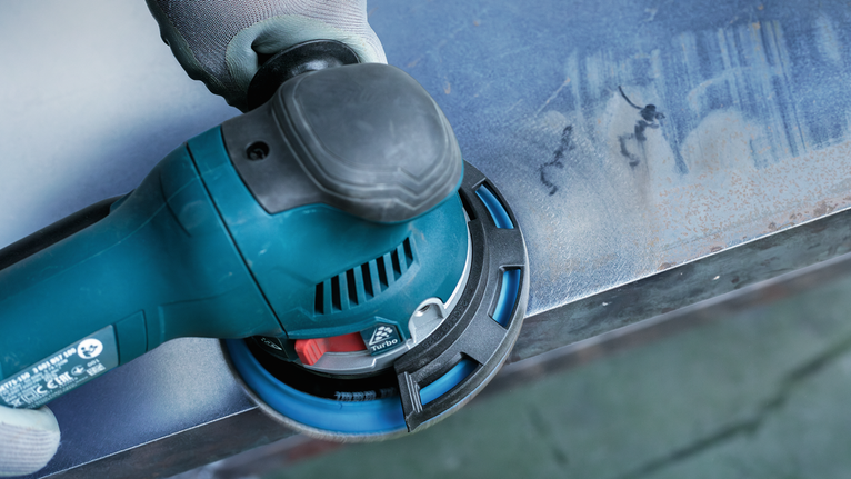 Person wearing safety equipment sanding a metal surface with a power tool.