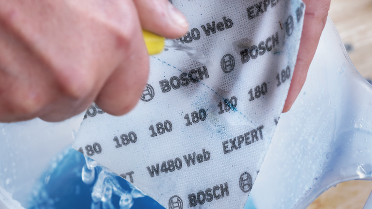 EXPERT W480 Web 1/4 Hand Sanding Sheet, 115 x 140 mm, G180 Person wearing safety equipment wets and scrubs abrasive sandpaper in a container.