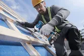 A person wearing safety equipment uses a cordless drill to fasten wood on a sloped surface.