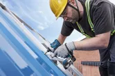 A person wearing safety equipment uses a cordless power tool to fasten screws on a metal roof.