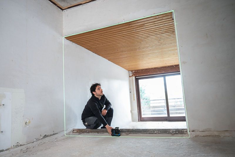 Person adjusting a laser leveling tool projects green lines in a partially finished room.