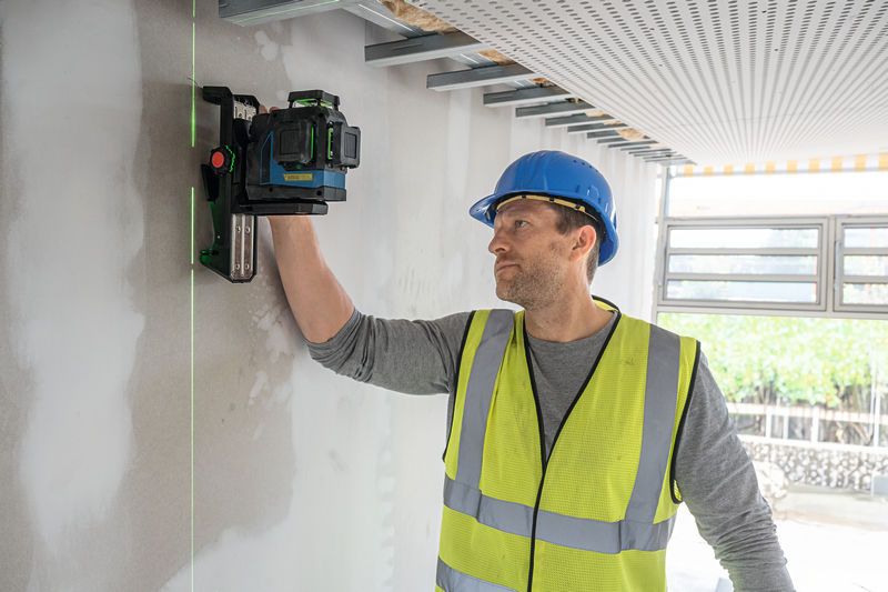 A person wearing safety equipment aligns a laser leveling tool on an unfinished wall.