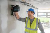 A person wearing safety equipment aligns a laser leveling tool on an unfinished wall.