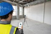 Person wearing safety equipment operates laser leveling tool remotely in an unfinished room.