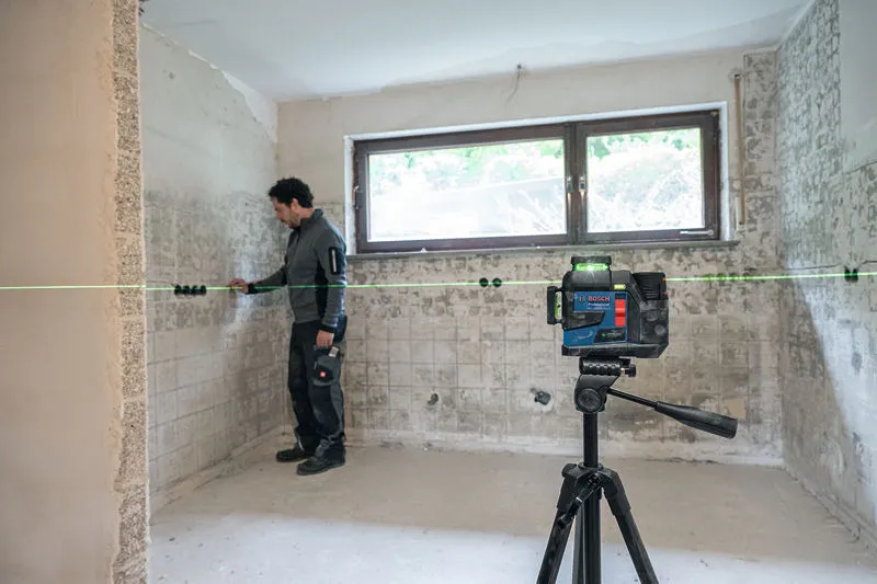 A person wearing safety equipment aligns wall sockets with a laser leveling tool in a tiled room.