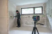 A person wearing safety equipment aligns wall sockets with a laser leveling tool in a tiled room.