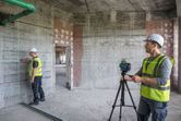 Two people wearing safety equipment use a laser leveling tool to align a wall in a construction site.