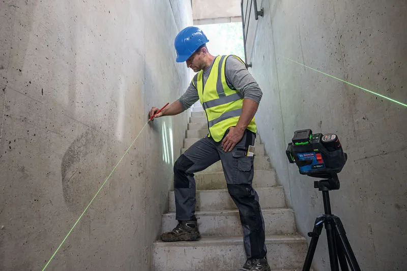 A person wearing safety equipment marks a line on a stairwell wall using a laser leveling tool.