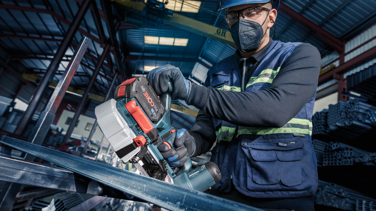 A person wearing safety equipment uses a cordless saw to cut metal beams.