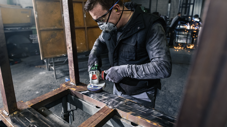 Person wearing safety equipment grinds metal on a workbench in an industrial setting.