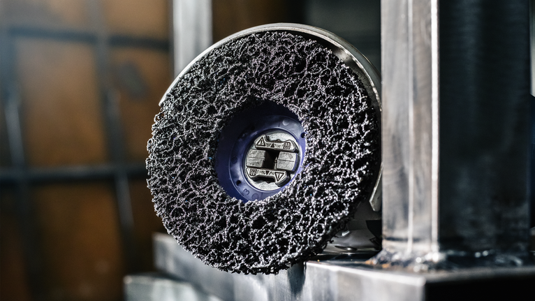 A coarse surface cleaning disc attached to a power tool rests on a metal structure.
