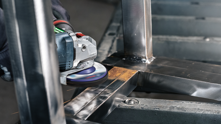 A person wearing safety equipment grinds a metal frame using an angle grinder.