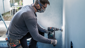 A person wearing safety equipment drills a hole in a wall with a power tool.