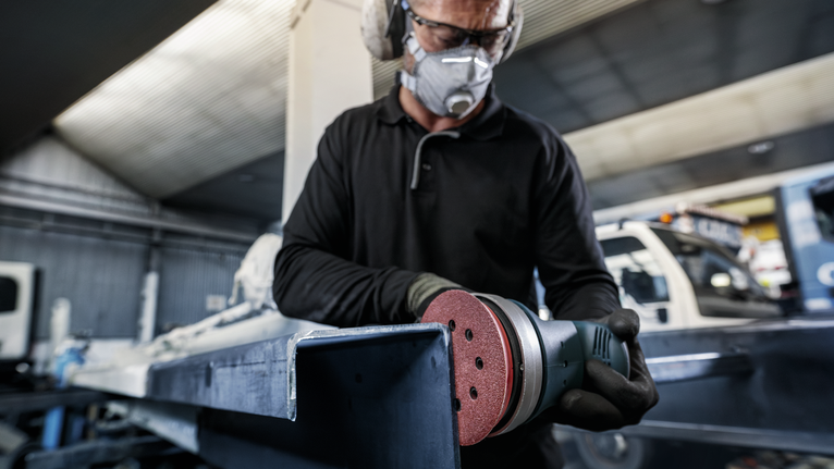 A person wearing safety equipment sands a metal beam with a power tool.