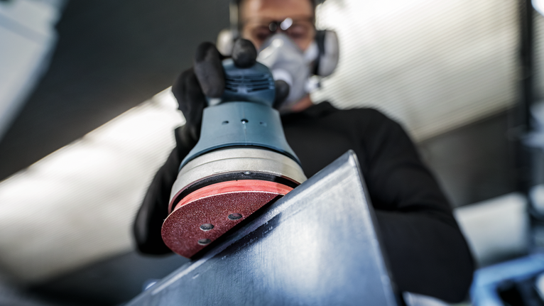Person wearing safety equipment sanding a metal surface with a power sander.