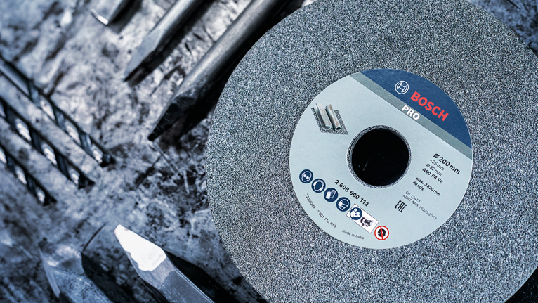 PRO Metal Grinding Wheel, 150 x 20 x 20 mm, G24 A grinding wheel lies on a workbench beside several metal drill bits.