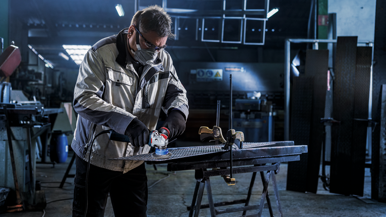 A person wearing safety equipment grinds a metal piece in an industrial workshop.