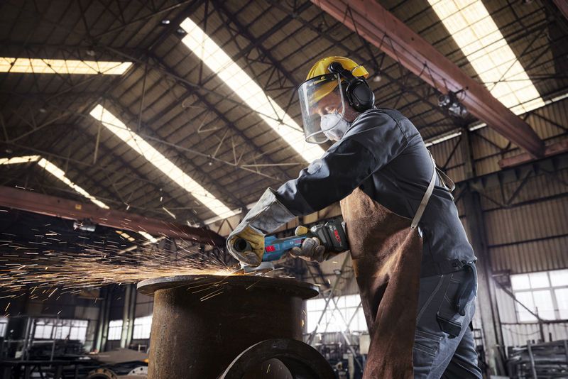 A person wearing safety equipment uses a cordless angle grinder to cut metal, creating sparks.