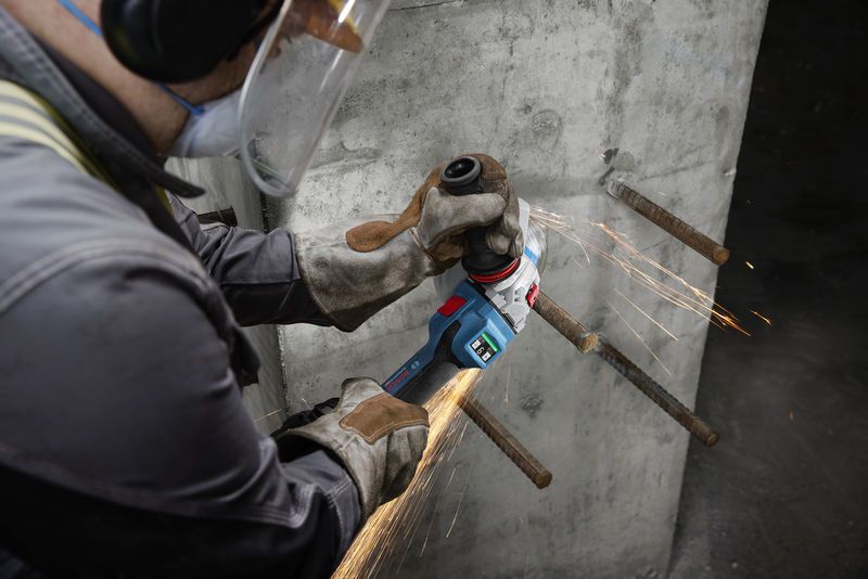 A person wearing safety equipment grinds exposed rebar on a concrete wall.