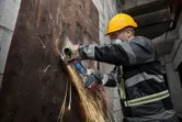 A person wearing safety equipment grinds a rusted metal sheet with a cordless angle grinder.