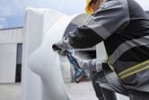 A person wearing safety equipment grinds a large metal pipe with a cordless angle grinder.