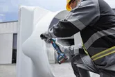 A person wearing safety equipment grinds a large metal pipe with a cordless angle grinder.