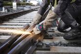 A person wearing safety equipment grinds a railway track with a cordless angle grinder.