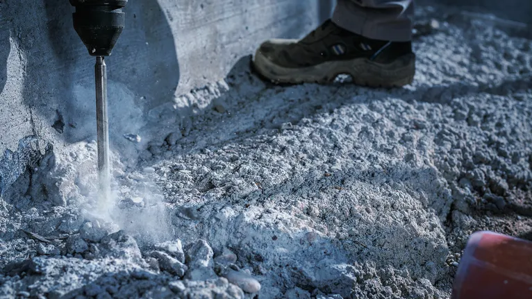 Person wearing safety equipment drills into concrete along a wall.