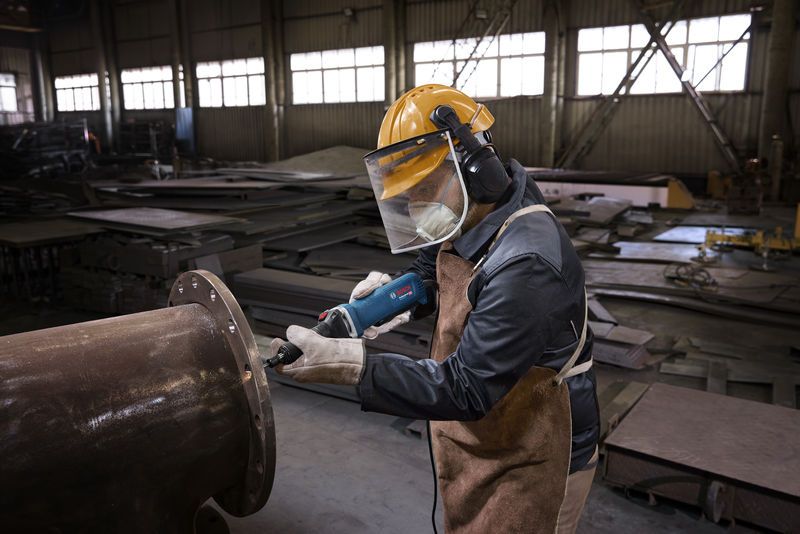 A person wearing safety equipment uses a straight grinder to work on a large metal pipe.