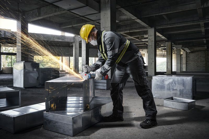 A person wearing safety equipment uses an angle grinder on a metal duct in a construction site.