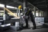 A person wearing safety equipment uses an angle grinder on a metal duct in a construction site.