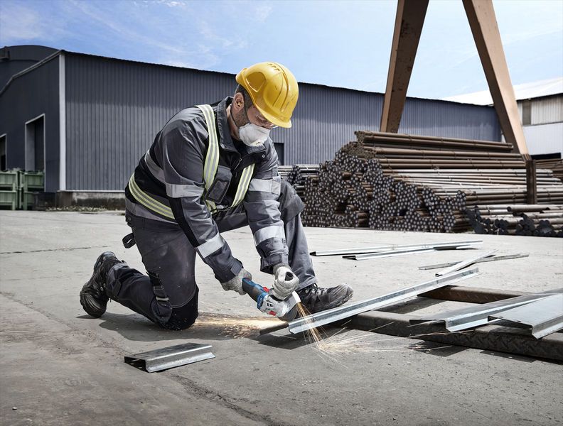 A worker wearing safety equipment cuts metal with a power grinder at an industrial site.