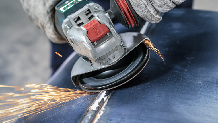 PRO Backing Pad Clip, X-Lock Person wearing safety equipment grinds a metal surface with an angle grinder, producing sparks.