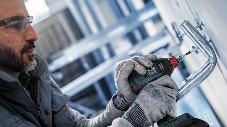 A person wearing safety equipment uses a power drill to fasten a bracket to a wall.