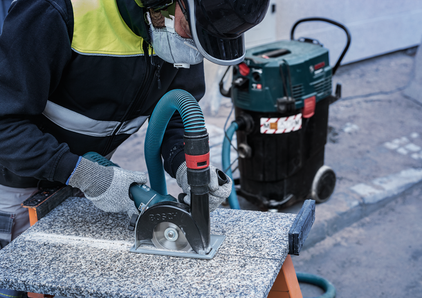Person wearing safety equipment cuts stone with a circular saw connected to a dust extractor.