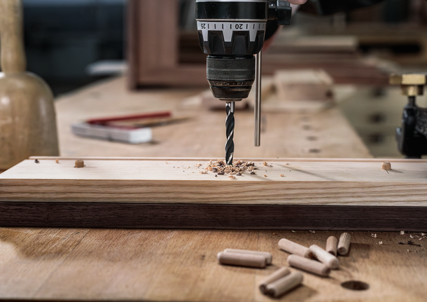 A person drills a hole into a wooden board on a workbench.