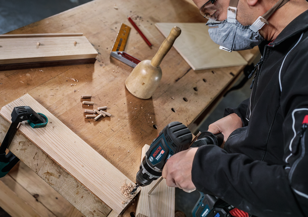 Person wearing safety equipment drills into wood on a workbench.