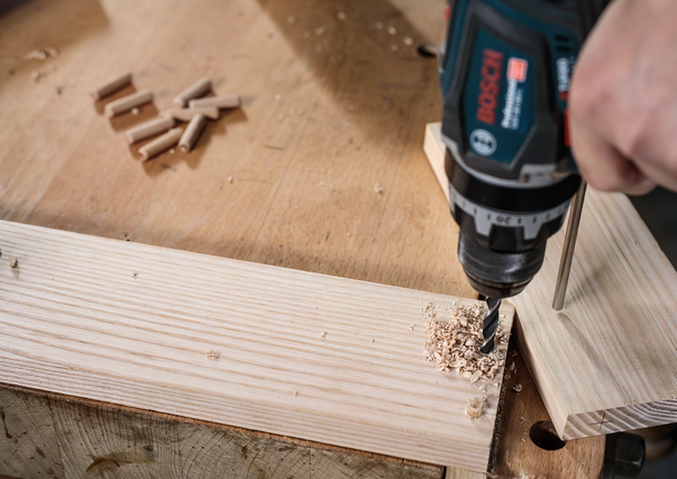 Person drills a hole in a wooden board on a workbench.