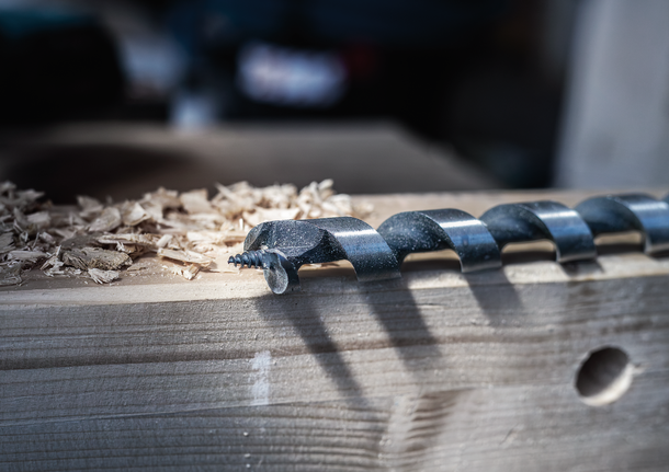 Large auger drill bit resting on a wooden plank with wood shavings beside it.