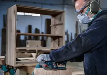 Person wearing safety equipment sands wooden furniture with a power tool.