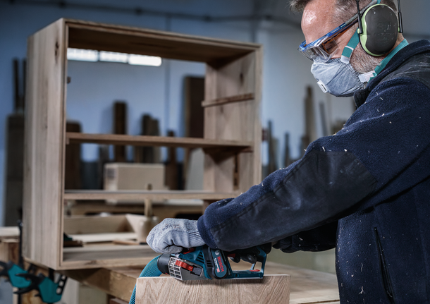 Person wearing safety equipment sands wooden furniture with a power tool.