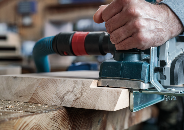 Person shapes the edge of a wooden board using an electric planer in a workshop.