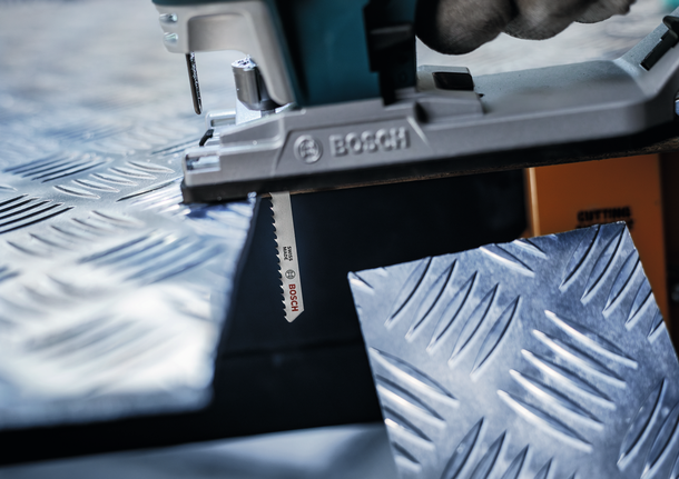 PRO Alu T127D Jigsaw Blade, 100 mm, 5 pcs Person cuts textured metal sheet with a jigsaw, wearing safety equipment.