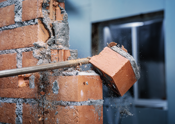PRO SDS max-4C Flat Chisel, 25 x 600 mm A demolition tool breaks a brick wall, scattering debris.